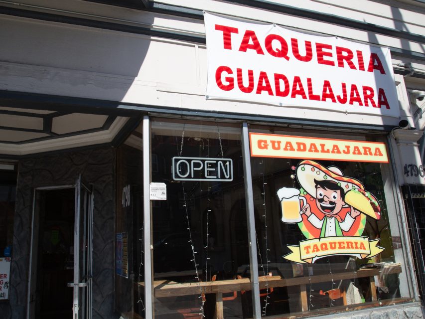 Storefront of Taqueria Guadalajara with a large sign, an "OPEN" neon light, and a cartoon character holding a taco and beer on the window.