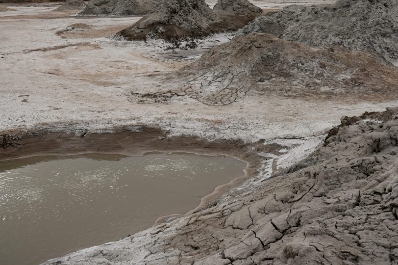 Cracked earth with small mounds and a pool of murky water in a dry, barren landscape.