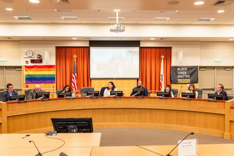 People sit around a semi-curcular dais, in front of a rainbow flag and flag reading "Black Lives Matter."