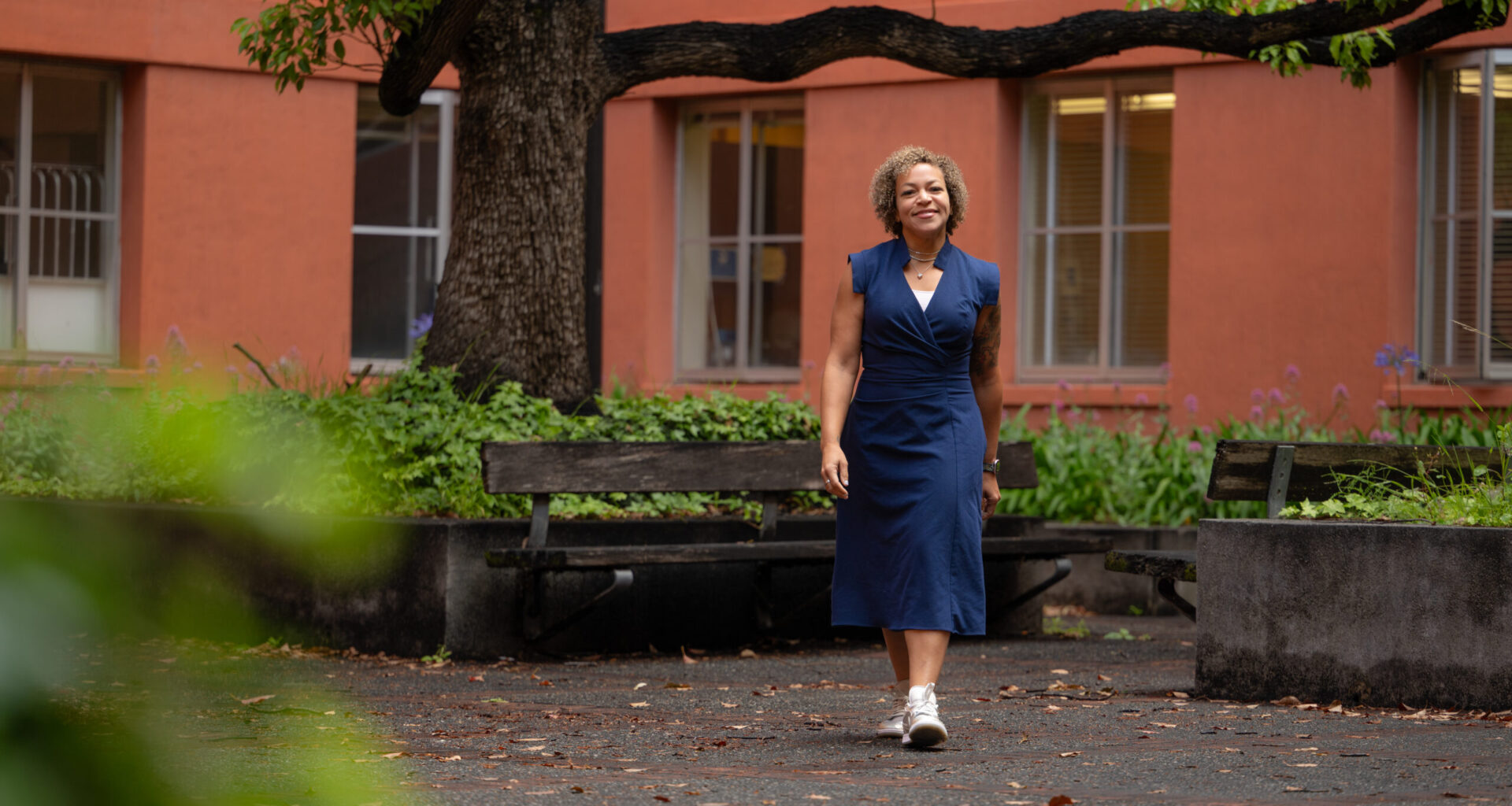 Nicole Holliday, in a navy sleeveless dress, walks along a tree-shaded path outside a salmon-colored building.
