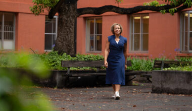Nicole Holliday, in a navy sleeveless dress, walks along a tree-shaded path outside a salmon-colored building.