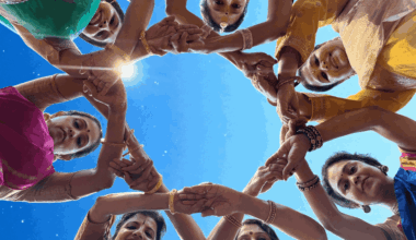 A group of women in colorful traditional attire stand in a circle, joining hands and looking down towards the camera against a clear blue sky.