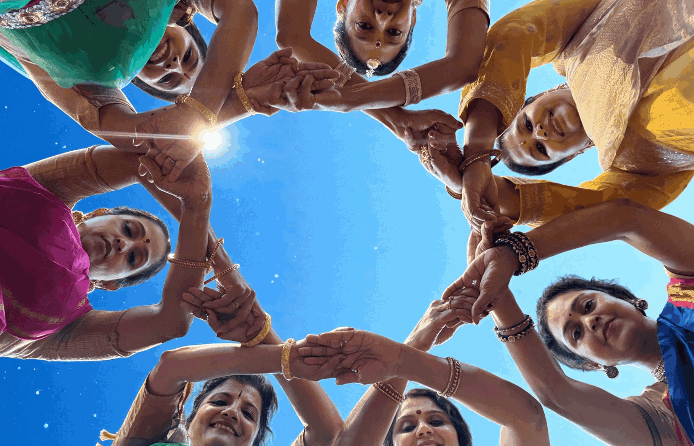 A group of women in colorful traditional attire stand in a circle, joining hands and looking down towards the camera against a clear blue sky.