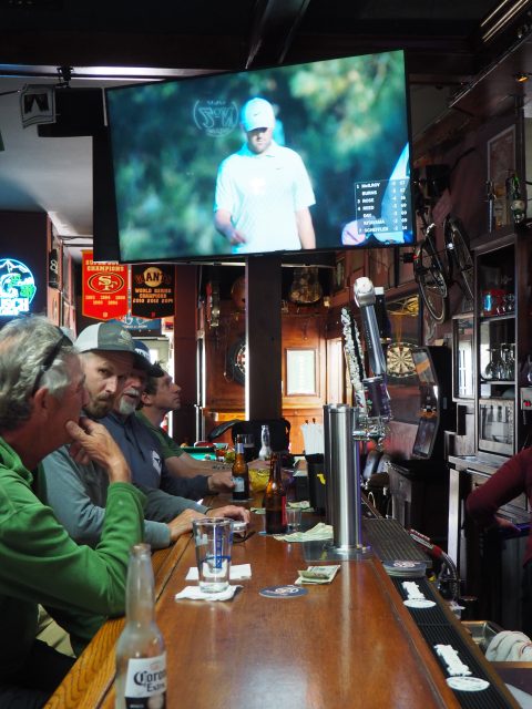 Four men sit at a bar on Noriega Street watching a golf tournament on a large TV above, with drinks and bottles on the counter in front of them.