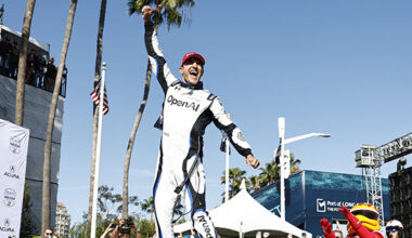 Race car winner on the podium raising a fist, wearing a white racing suit with OpenAI logo, palm trees and blue sky behind.