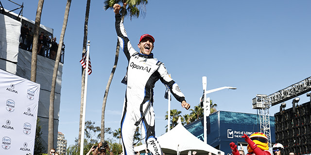 Race car winner on the podium raising a fist, wearing a white racing suit with OpenAI logo, palm trees and blue sky behind.