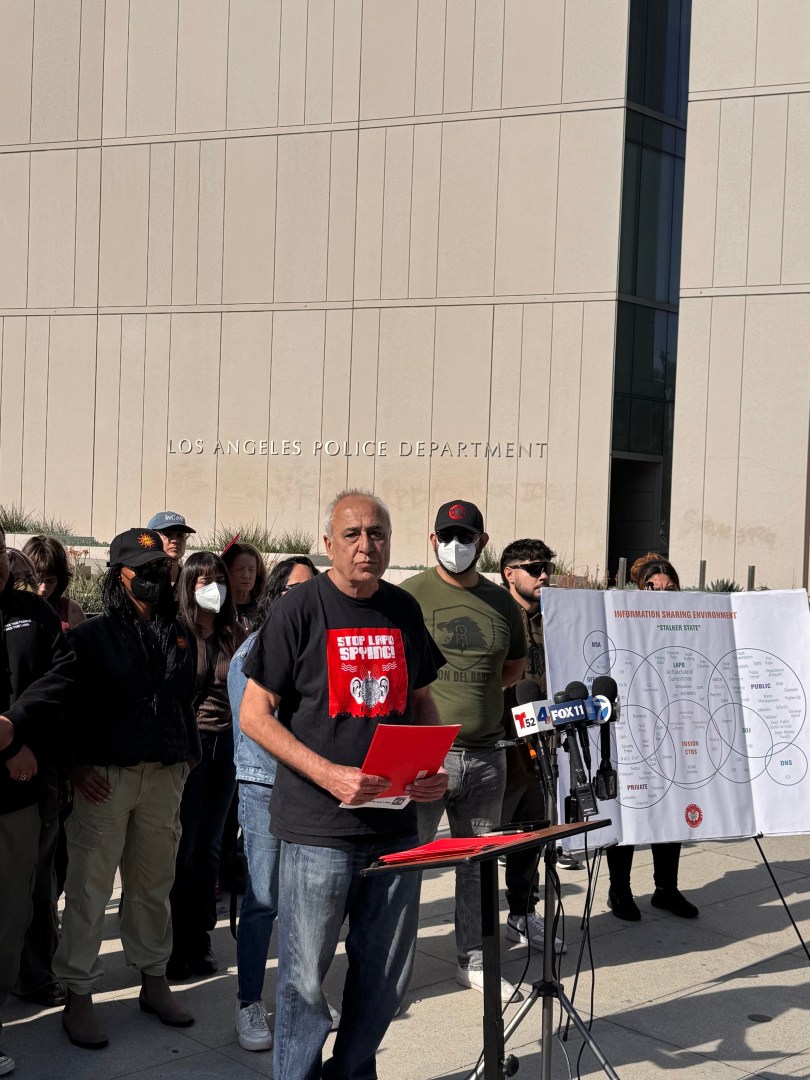 A photo of a man in a "Stop LAPD Spying!" shirt speaking at a podium with a crowd behind him in front of the Los Angeles Police Department.
