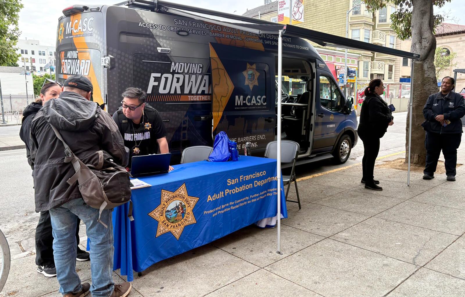 A San Francisco Adult Probation Department mobile van is parked on a city street with staff assisting people at an outdoor table covered by a branded tablecloth.