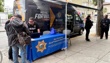 A San Francisco Adult Probation Department mobile van is parked on a city street with staff assisting people at an outdoor table covered by a branded tablecloth.