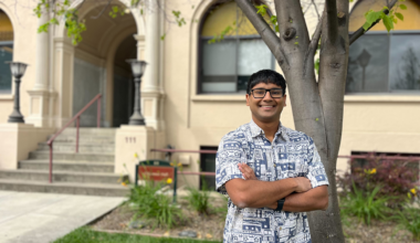 A young man in a patterned shirt stands arms crossed by a tree in front of an academic building.