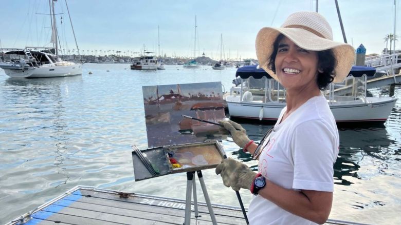 Woman smiling while painting on a dock overlooking a harbor