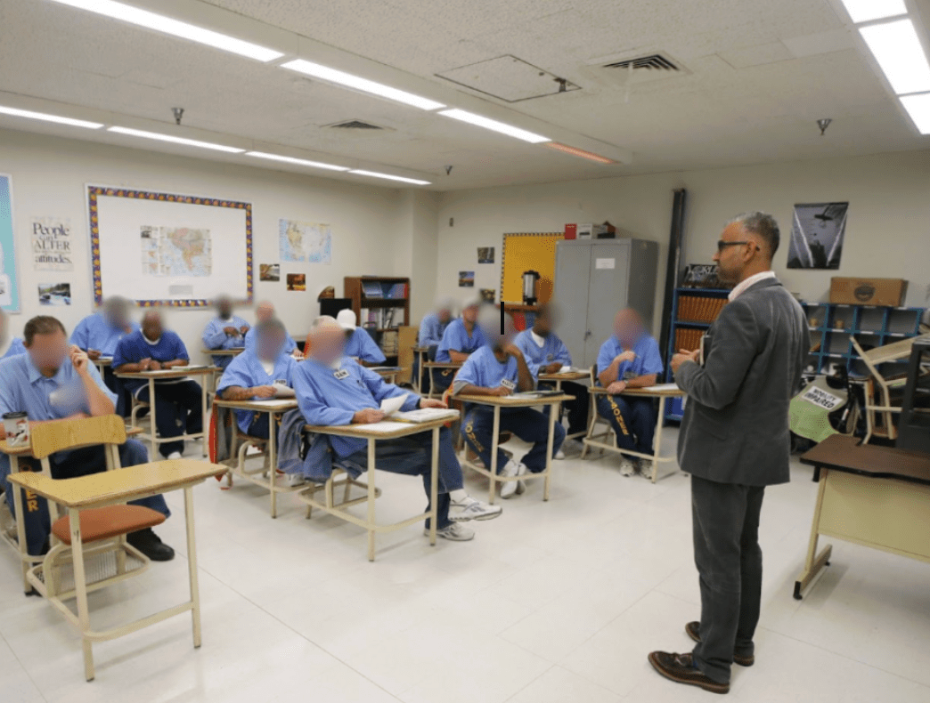 A photo of a man in suit standing in front of a incarcerated men, whose faces are blurred, sitting in desks in a classroom.