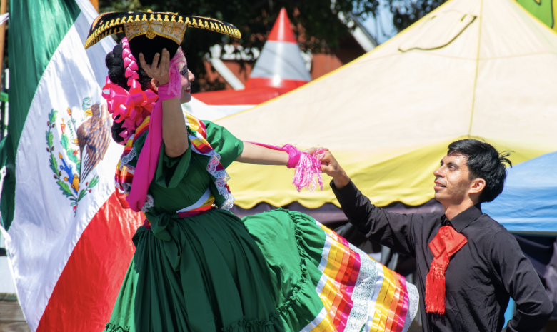 A man and woman perform a traditional Mexican dance. The woman wears a vibrant green dress and sombrero, and the man is dressed in black with a red scarf. A Mexican flag waves behind them, adding to the festive atmosphere.