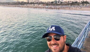Man wearing sunglasses and an LA cap takes a selfie on a pier overlooking a crowded beach and city buildings in the background at sunset.