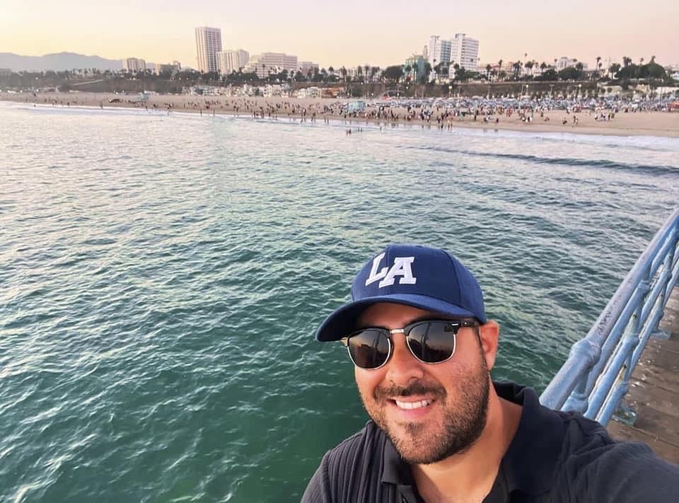 Man wearing sunglasses and an LA cap takes a selfie on a pier overlooking a crowded beach and city buildings in the background at sunset.