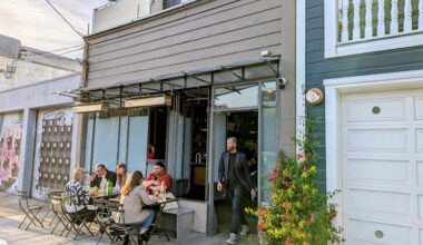 People sitting at outdoor café tables and talking while a man exits the building; plants and a garage are visible beside the café.