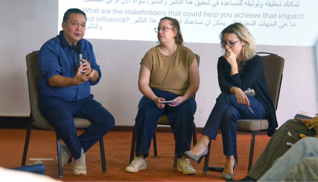 A seated man at left holds a microphone and is talking, while two women sitting next to him listen intently. Behind them is a slide projection in English and Arabic.