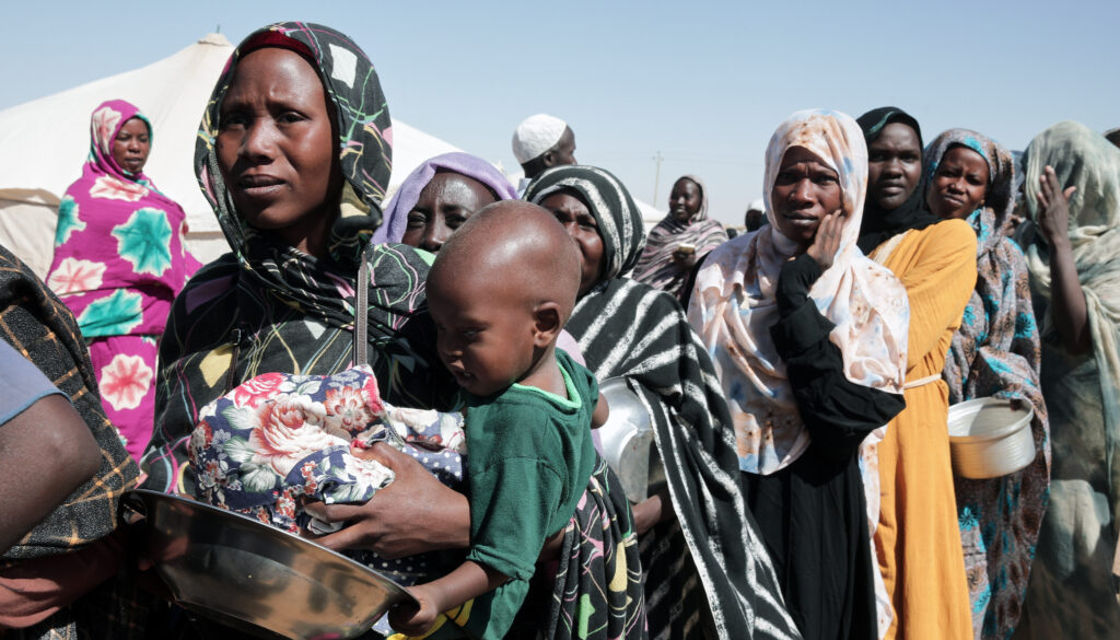 Solemn-looking Sudanese women refugees wearing robes in bold prints and bright colors wait for food under the desert sun. The woman at the front of the line is holding a young child and a metal bowl.
