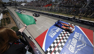Race cars cross the finish line at a Grand Prix as a green flag is waved from the trackside grandstand.
