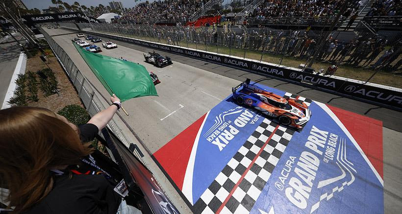 Race cars cross the finish line at a Grand Prix as a green flag is waved from the trackside grandstand.