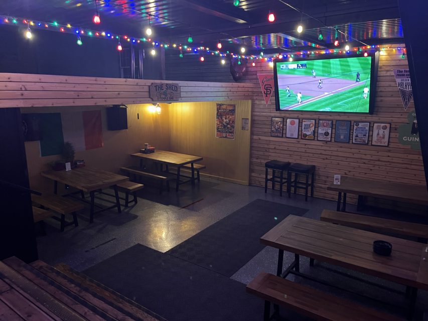 A dimly lit bar with wooden benches and tables, colorful string lights overhead, and a TV on the wall showing a baseball game.