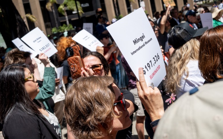 People holding signs that say "Mingei International Museum: $197,326 eliminated" amid a crowd of San Diegans who turned out on April 20, 2026 to oppose city budget cuts affecting the arts. (Photo by Thomas Murphy/Times of San Diego)