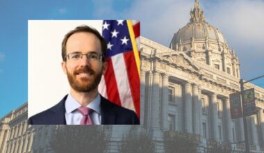 A man in a suit and tie stands in front of an American flag; in the background is a large government building with a dome and ornate architecture.