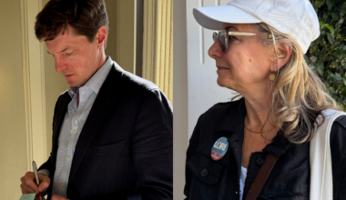 A man in a suit writes on a clipboard indoors, while a woman in a white cap and glasses stands outside, wearing a "LORI" campaign button.