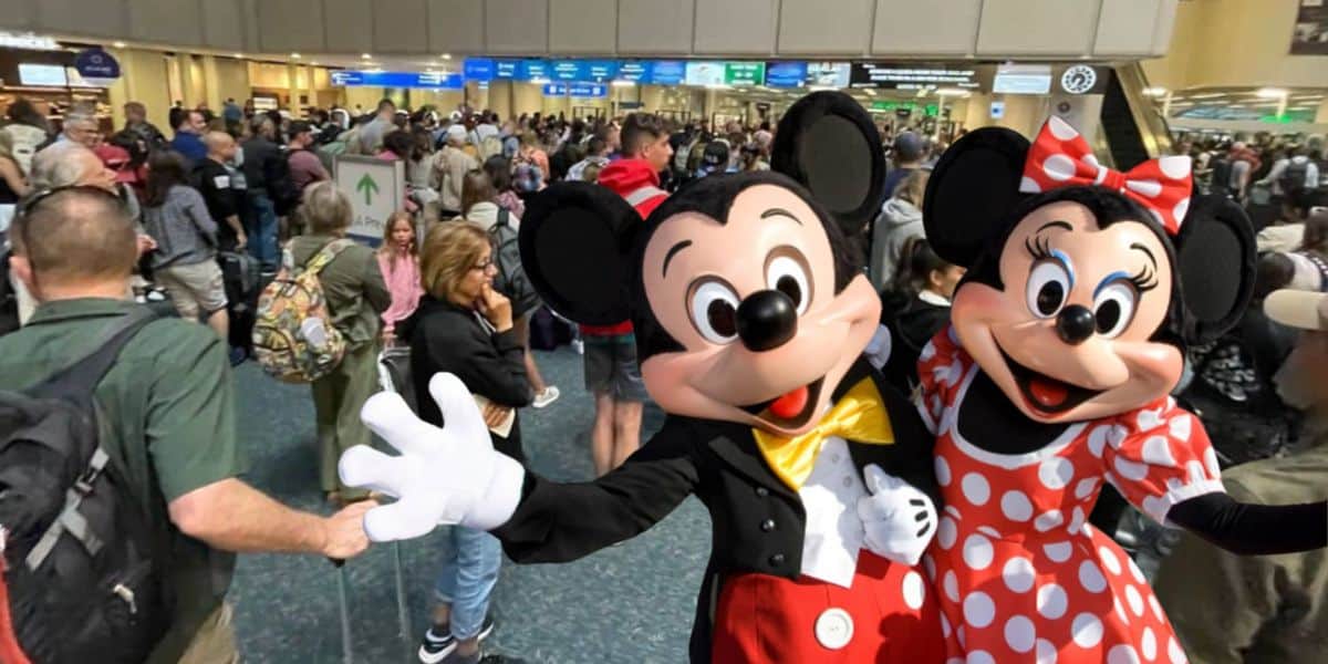 Mickey Mouse and Minnie Mouse greet and pose for a photo in a crowded Orlando International Airport terminal, surrounded by travelers and people with luggage as Disney guests get United Airlines 5% of their flights cut due to rising fuel costs.