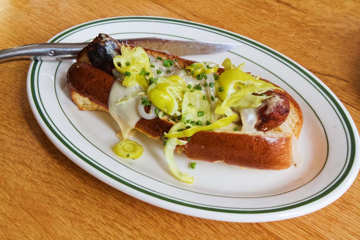 A horizontal photo of a sausage hot dog smothered in cheese and pepperoncini. A French steak knife rests on the plate