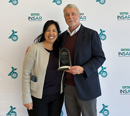 From left, Christine Wu Nordahl and David Amaral stand together, Amaral holding a glass award, in front of a white fabric backdrop that says INSAR