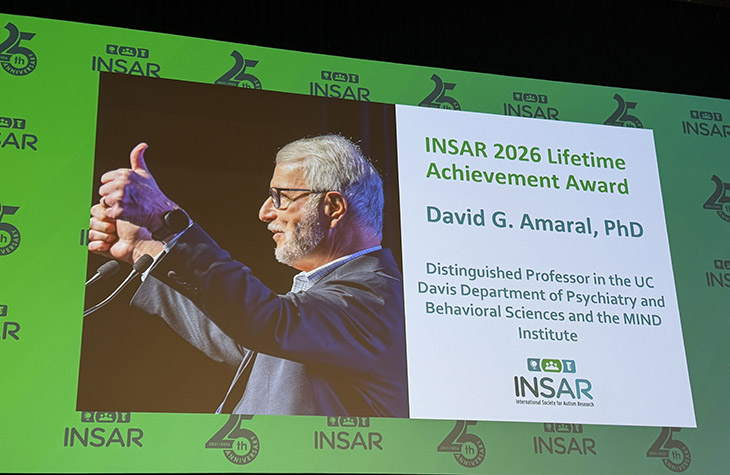 A large video screen above a stage shows David Amaral doing a thumbs up sign with text that reads INSAR Lifetime Achievement Award 2026,UC Davis distinguished professor of psychiatry and behavioral sciences and MIND Institute with the INSAR logo