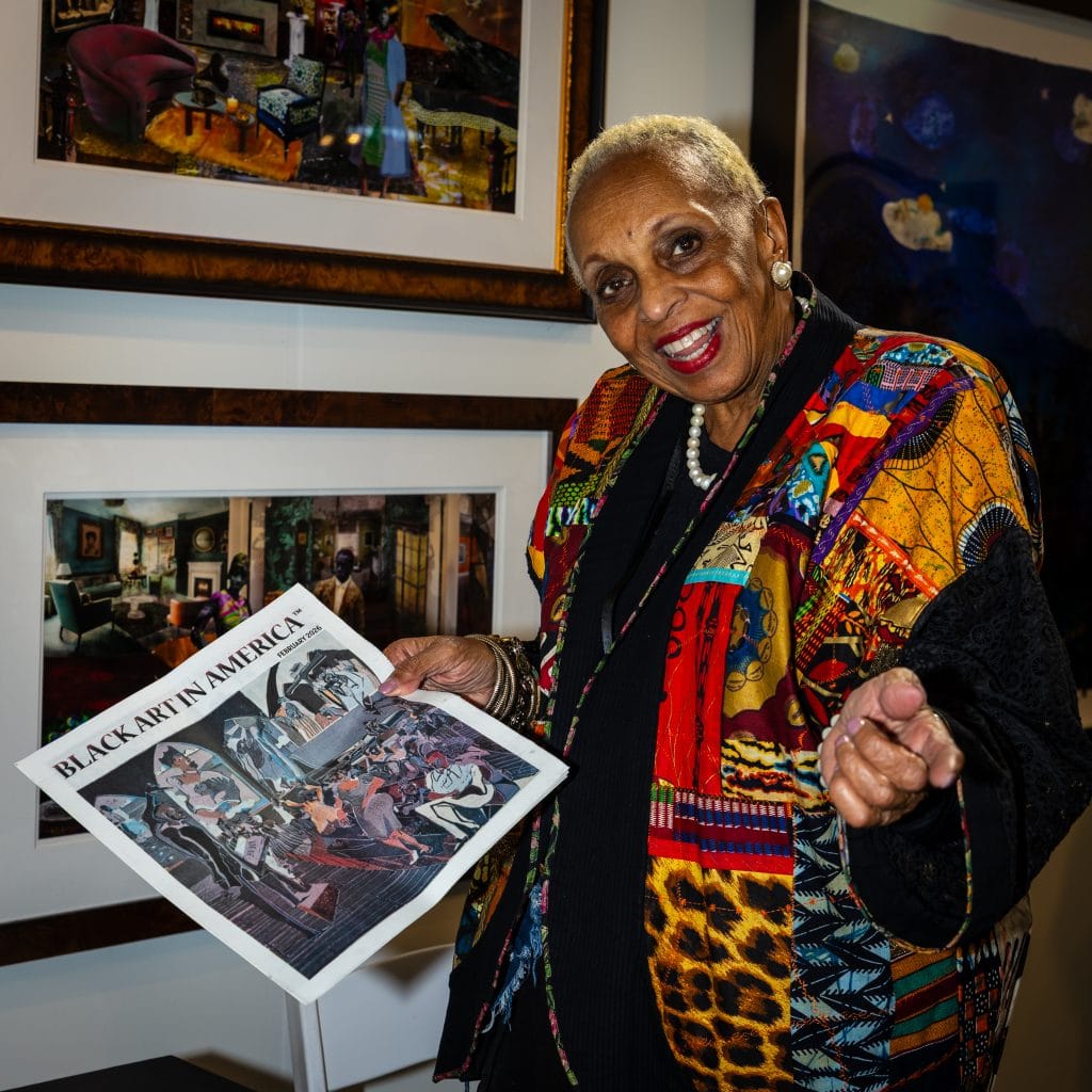An woman with short gray hair smiles warmly at the camera while holding a newspaper titled “Black Art in America.” She wears a colorful patterned jacket, pearl earrings, and a pearl necklace. Behind her, framed artworks hang on a gallery wall, featuring richly colored interior scenes and figures, suggesting an art exhibition setting. From the premier of San Francisco Art Fair.