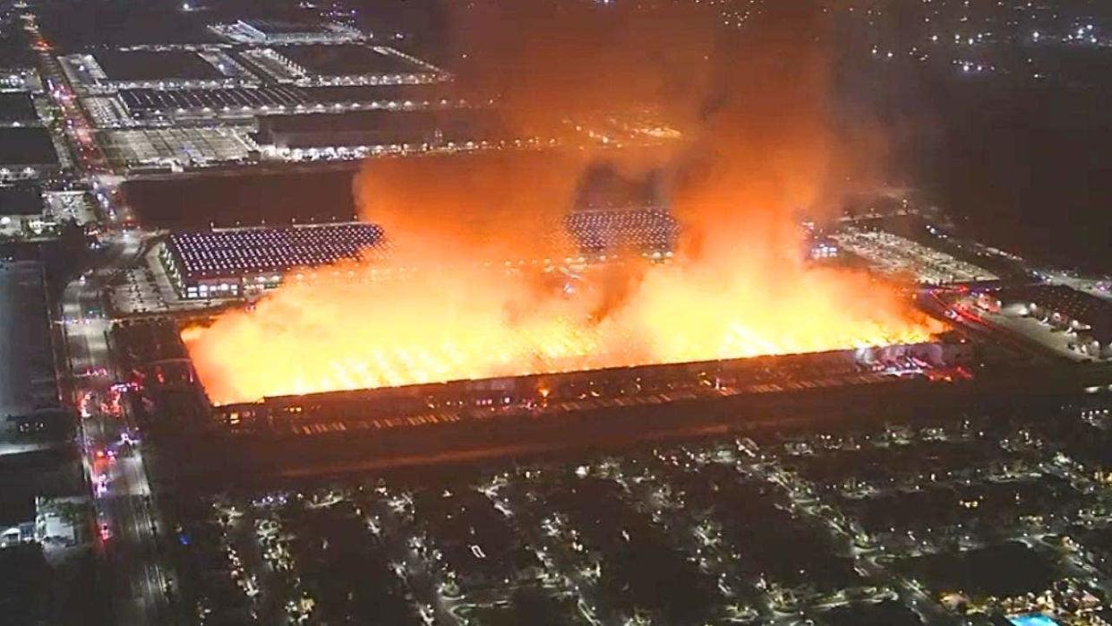Aerial nighttime image of a large warehouse fully engulfed in flames with thick smoke rising over an industrial area.