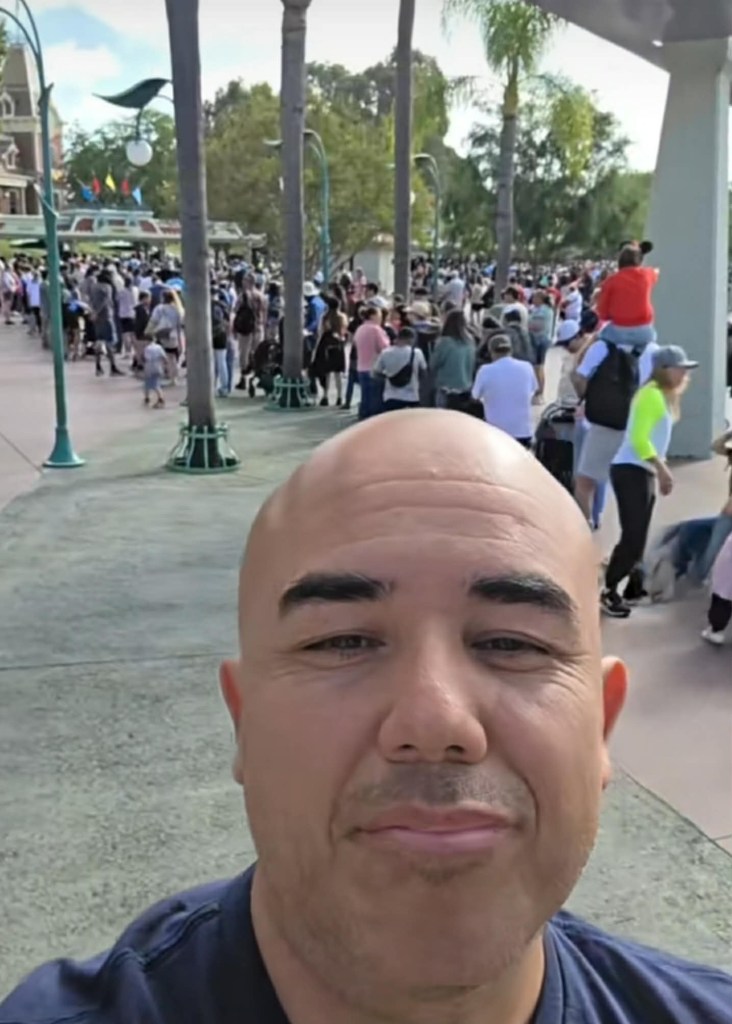 Nef, a bald man, taking a selfie in front of a crowd at Disneyland.
