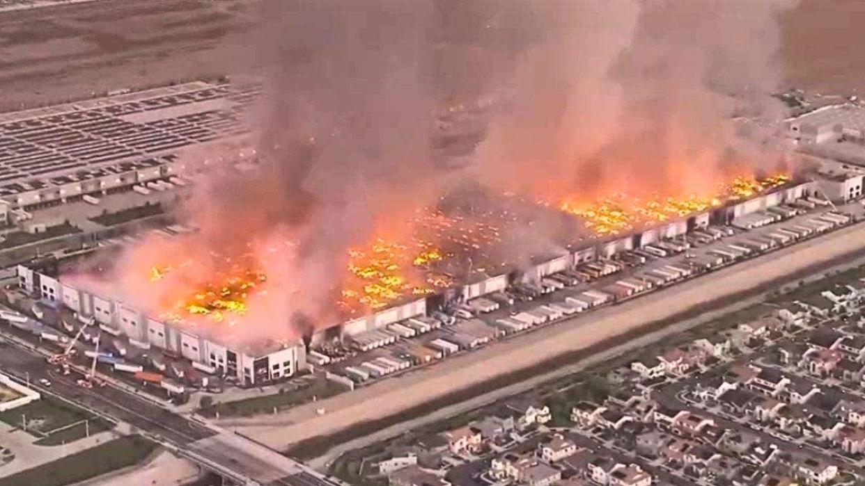 Aerial view of a large warehouse fire with flames and thick smoke rising over an industrial building in Ontario, California.