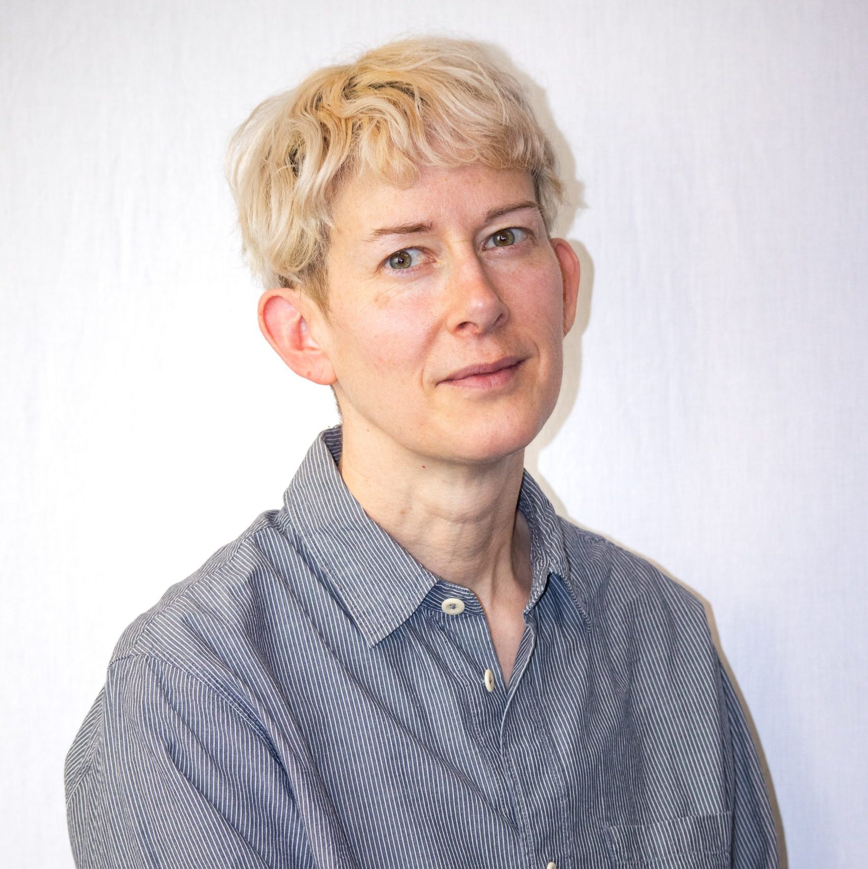 Person with short, light blonde hair wearing a striped button-up shirt, looking at the camera with a neutral expression against a plain white background.