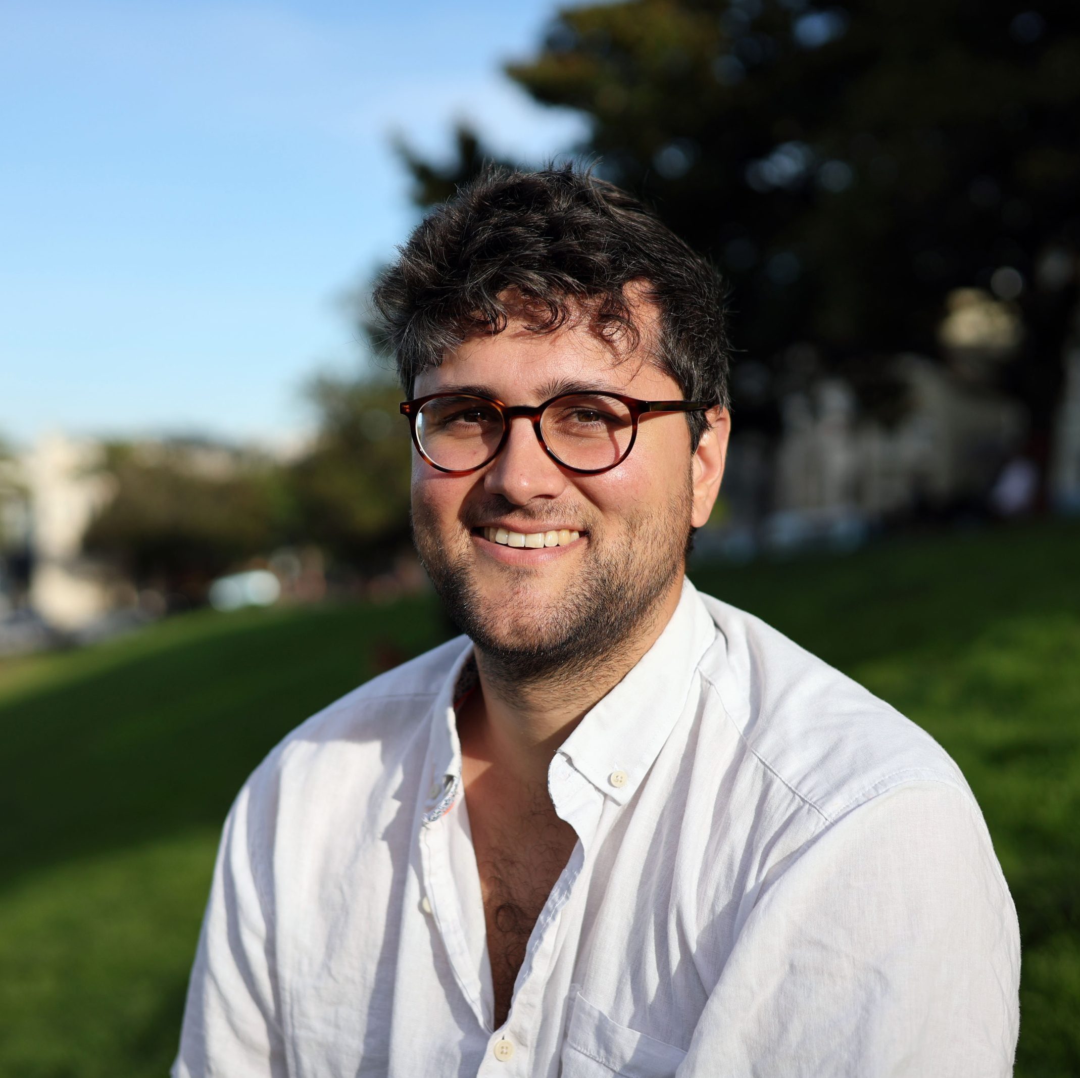 A man with dark hair and glasses, wearing a white shirt, sits on grass in a park on a sunny day, smiling at the camera.