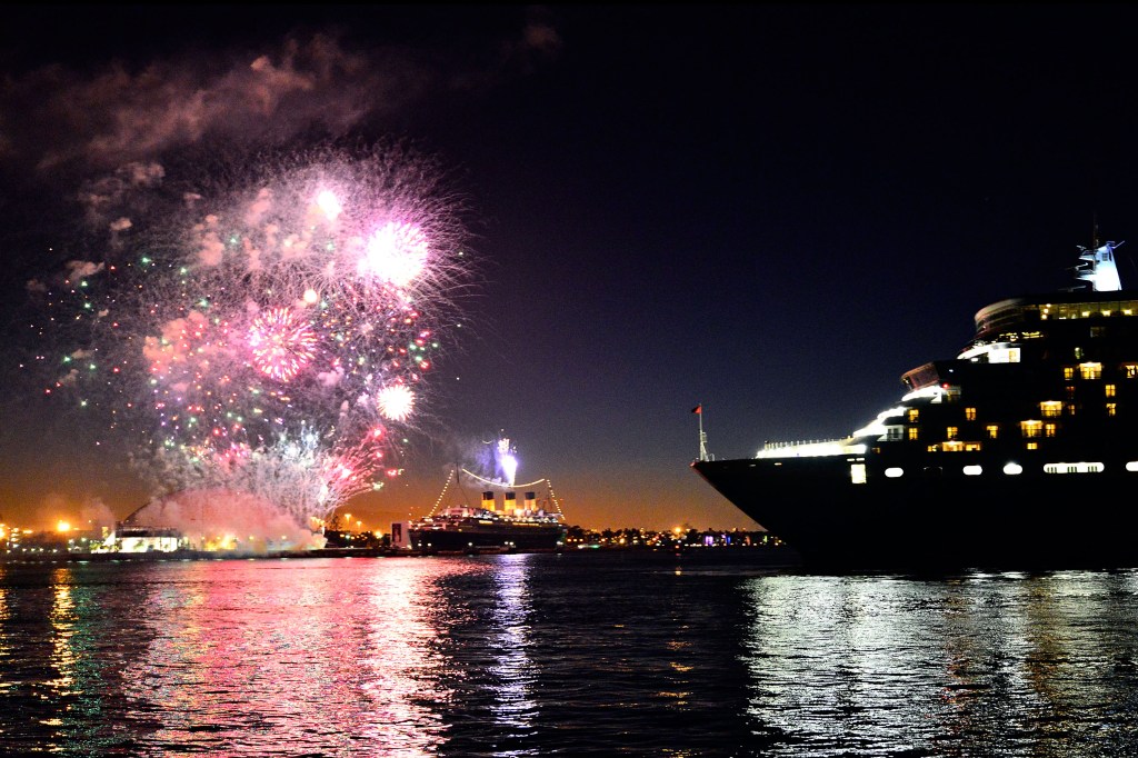 Cunard's Queen Elizabeth and Queen Mary cruise ships in Long Beach Harbor at night with fireworks in the sky.