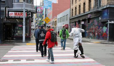 Amidst the vibrant cityscape, people cross the street, one draped in reflective material, reminiscent of a triage blanket. The backdrop of colorful buildings and dynamic street art adds an artistic flair to the urban scene.