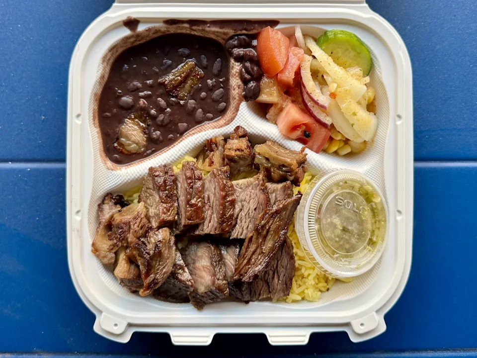 A plastic takeout box filled with beans, salad, and rice topped with asado, pork belly, and sausage.
