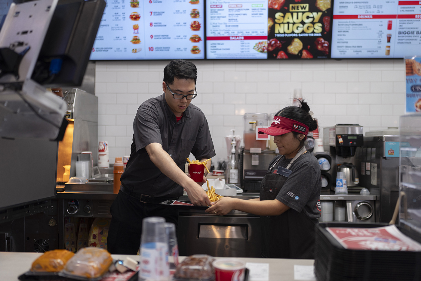 Two fast food workers in grey polos and black pants filling a Wendy's bag with food at a Wendy's location.