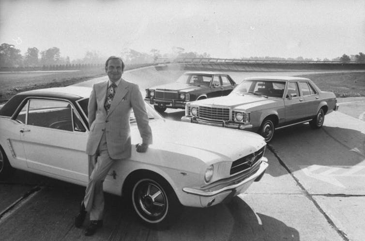 A man leans on a 1970s-ear car with two more behind him.