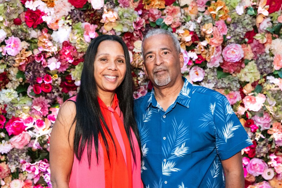 A woman and a man stand together in front of a colorful floral wall, both smiling at the camera.