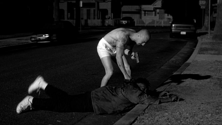 a man in underwear holding a gun beats up another man who is on the ground on the street
