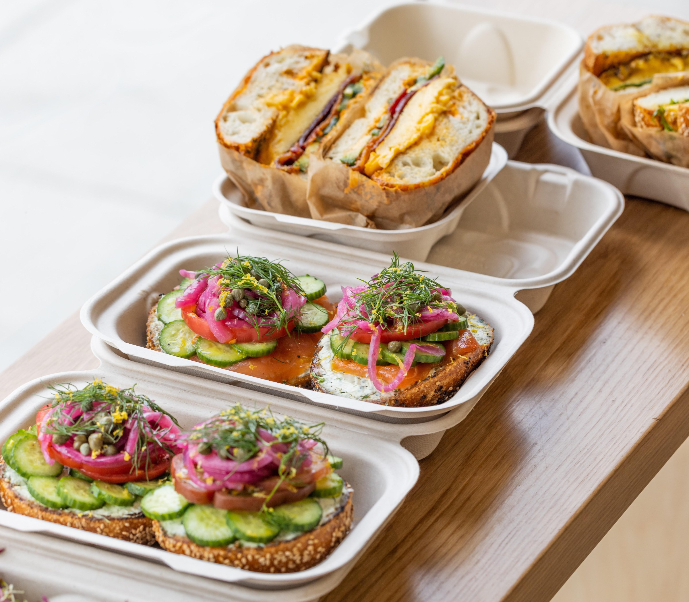 A sunny sidebar made of wood holding takeout boxes of bagels with vegetables, lox, and eggs and cheese at Layla Bagels in Santa Monica.