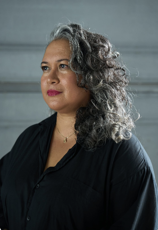A portrait of a woman with shoulder-length, curly gray hair, wearing a black blouse and a small gold necklace. She is looking off to the side with a calm, thoughtful expression. The background is softly lit and neutral, with subtle horizontal lines.