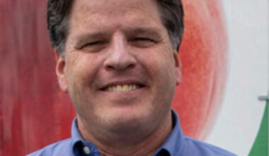 A man with dark hair wearing a blue collared shirt smiles at the camera, standing in front of the Los Angeles Regional Food Bank. A blurred image of a large fruit can be seen in the background, highlighting his commitment to nourishing communities.