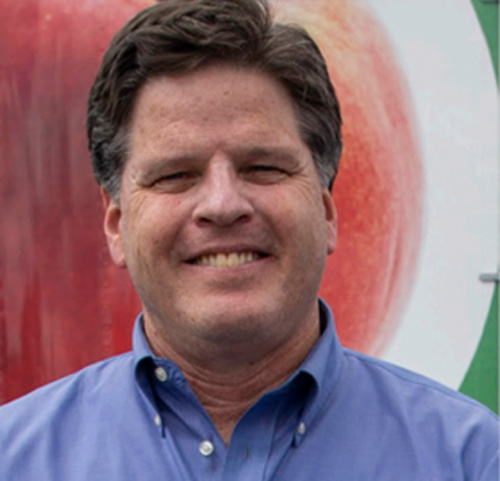 A man with dark hair wearing a blue collared shirt smiles at the camera, standing in front of the Los Angeles Regional Food Bank. A blurred image of a large fruit can be seen in the background, highlighting his commitment to nourishing communities.