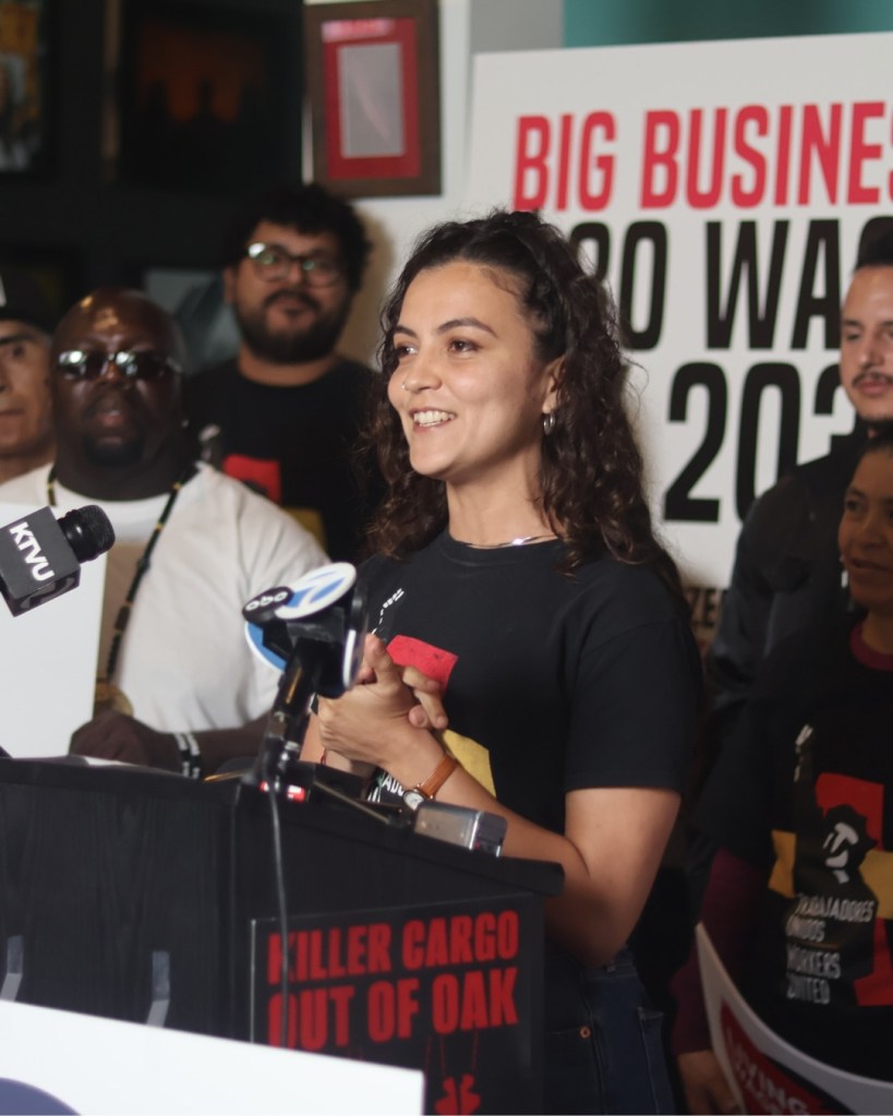 A smiling woman with curly hair speaks at a podium with a sign in the background reading "BIG BUSINESS" and "KILLER CARGO OUT OF OAK".